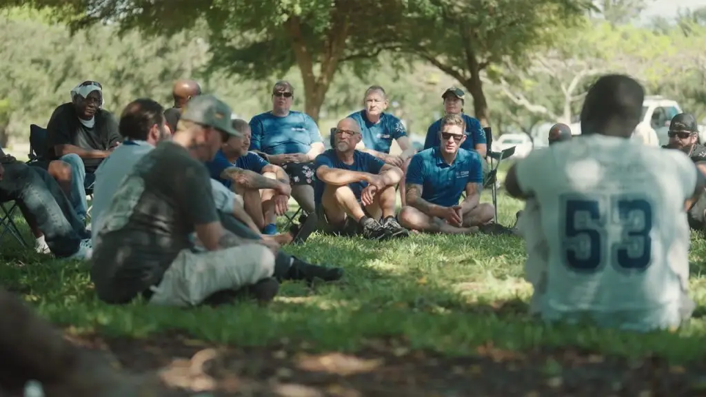 A diverse group of people from First Step Behavioral Health sit in a circle outdoors on grass under trees, engaged in a discussion. The atmosphere is relaxed and focused.
